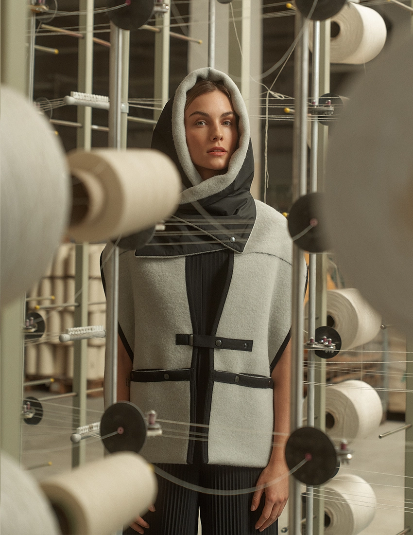 A model poses in a grey wool pocket scarf with a black high collar and straps, paired with a black ensemble, in a textile factory setting.