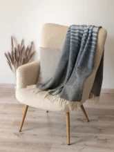 Recycled wool blanket in grey with soft blue lines, gracefully hanging on a contemporary cream armchair in a light-filled room.