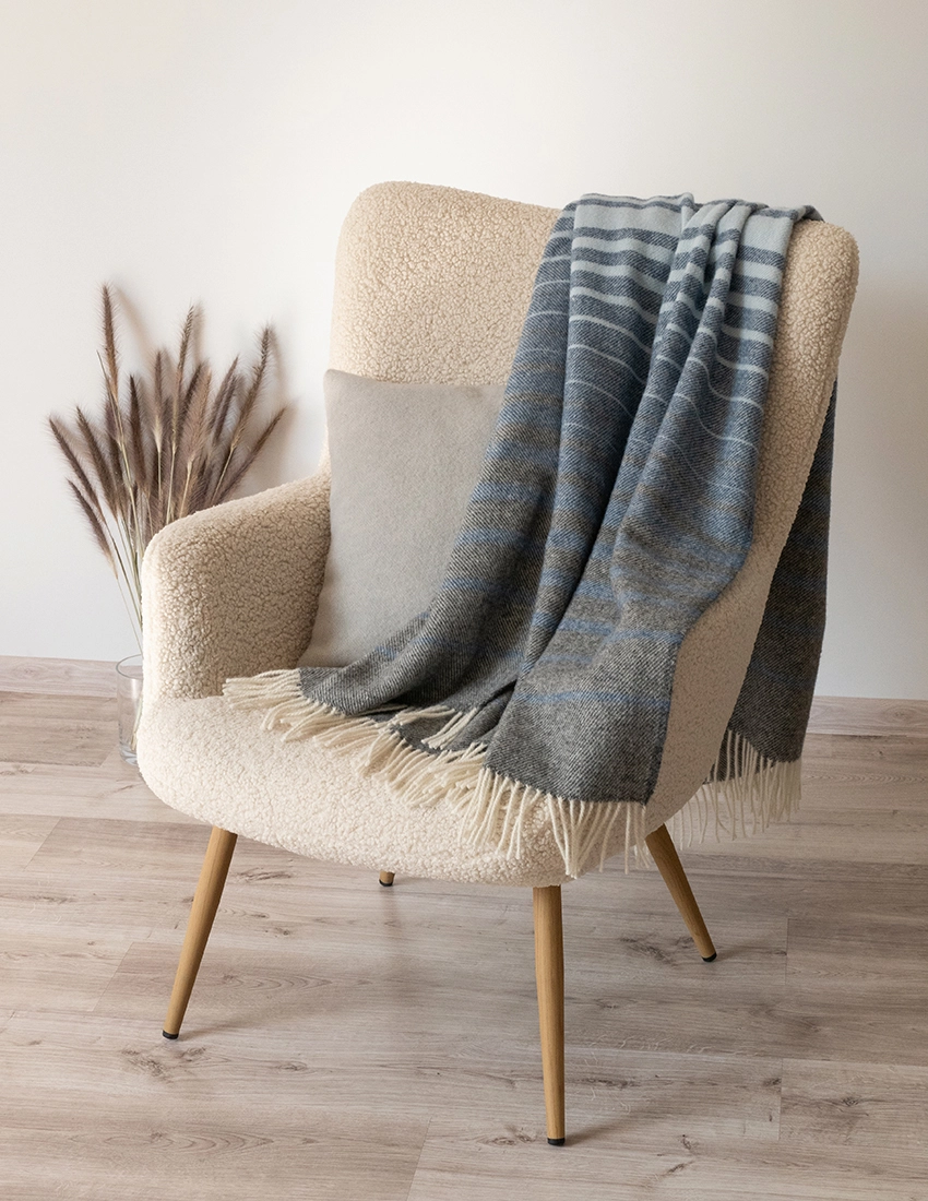 Recycled wool blanket in grey with soft blue lines, gracefully hanging on a contemporary cream armchair in a light-filled room.