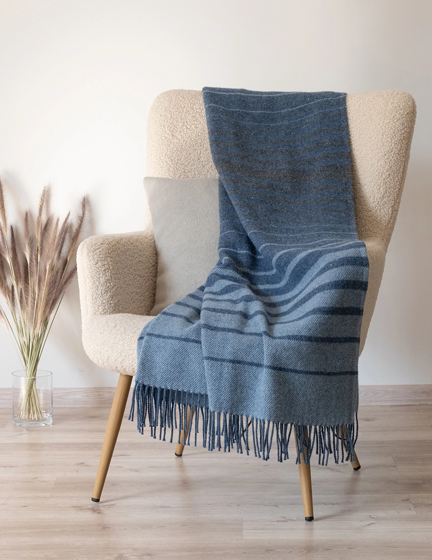 A cozy recycled wool blanket in shades of blue draped over a modern chair in a home setting, complementing the neutral room palette.